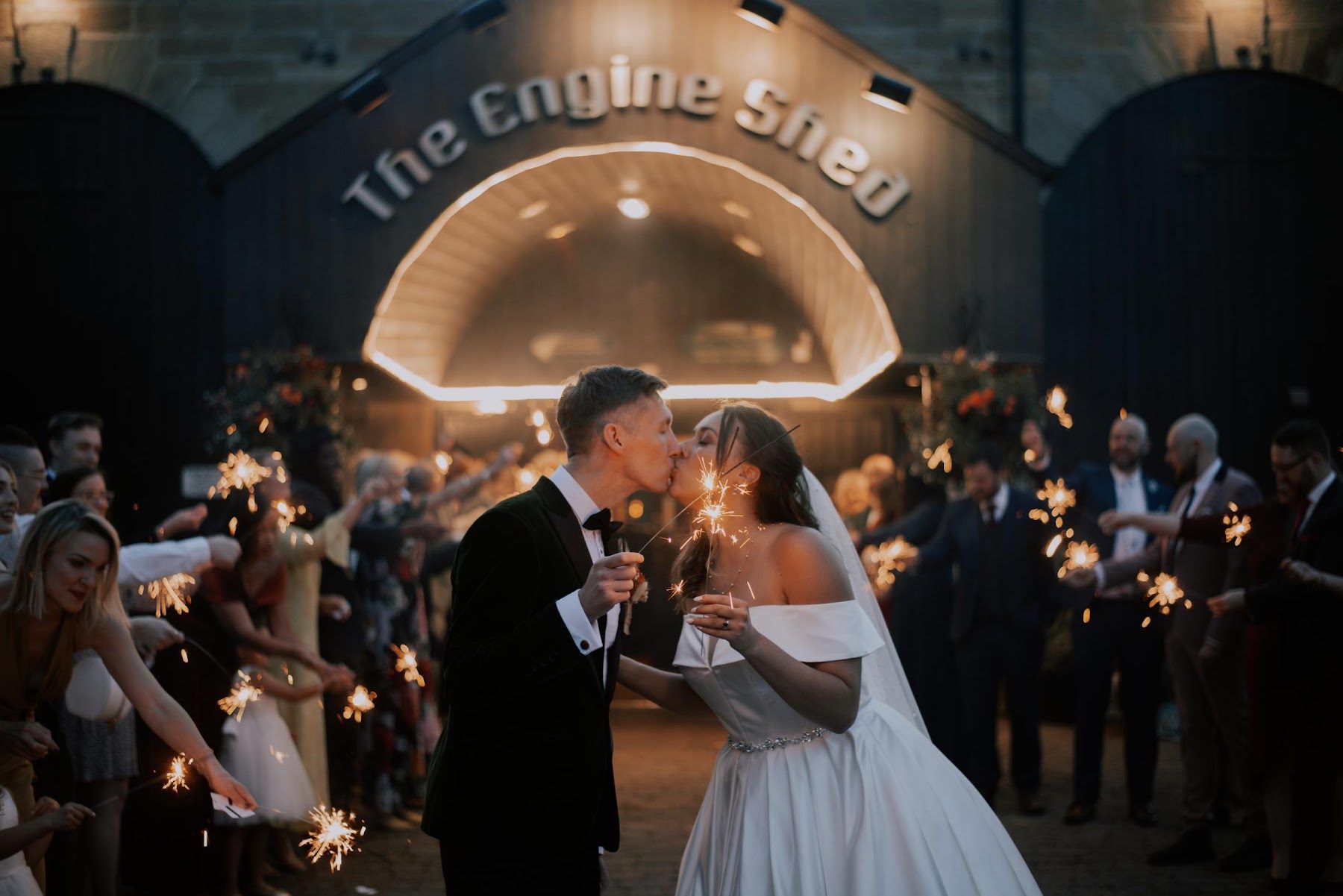 A couple kissing beneath the illuminated Engine Shed arch as their guests hold sparklers